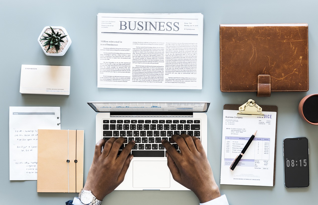 Photo of a man using a laptop, surrounded by a newspaper, planner, and clipboard.