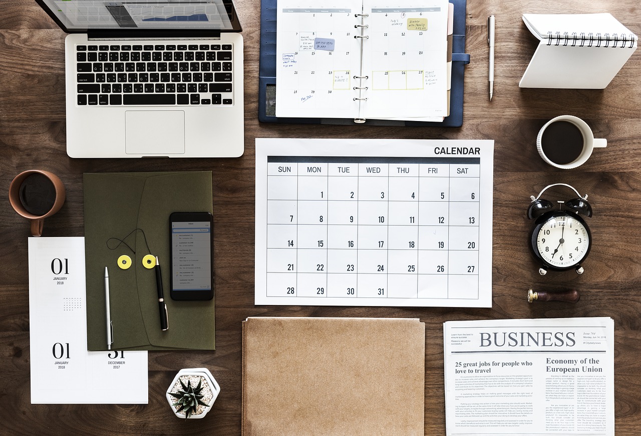 Photo of a desk with a laptop, calendar, planner, clock, and newspaper.