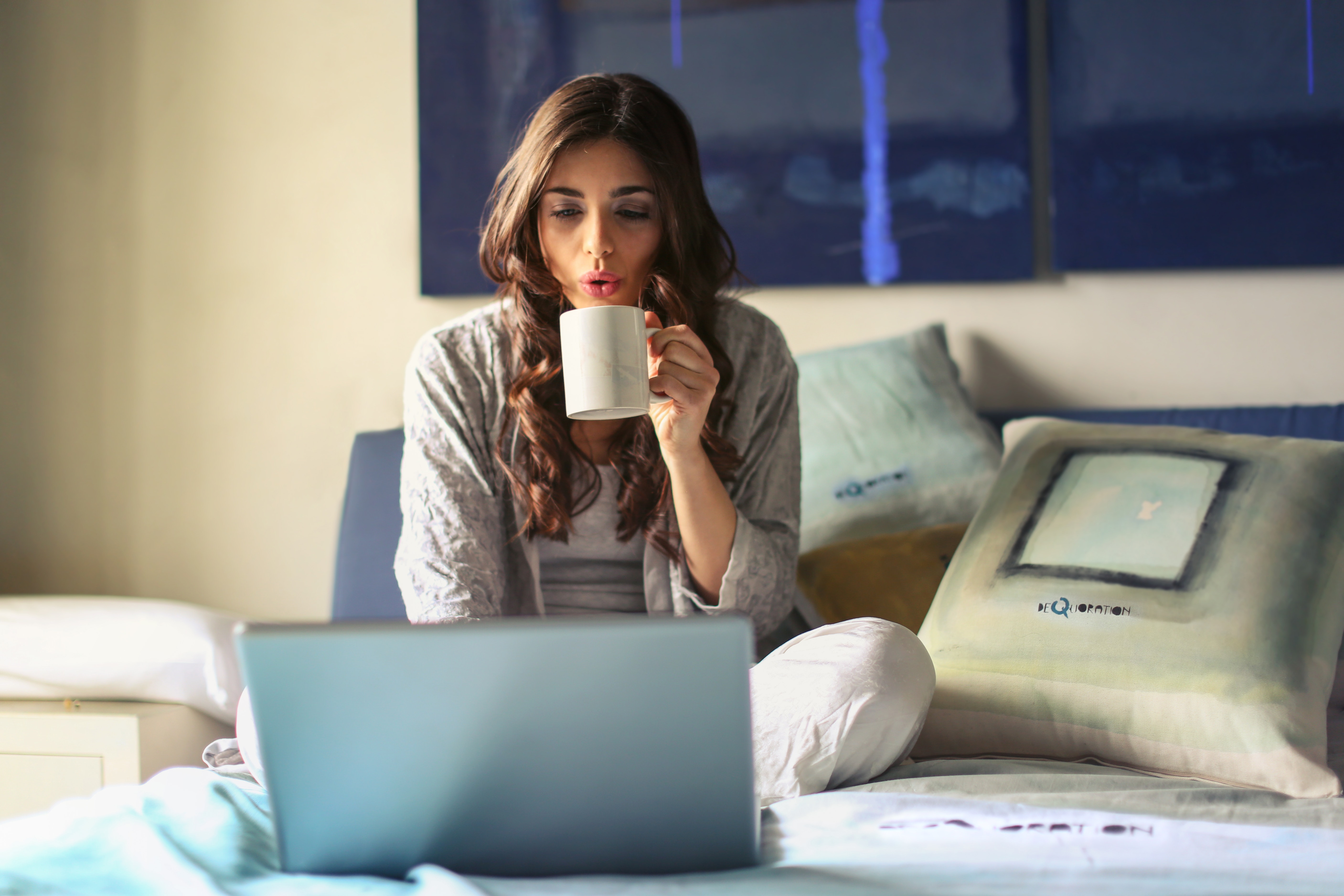 Photo of a woman drinking a cup of tea while using her laptop in bed.