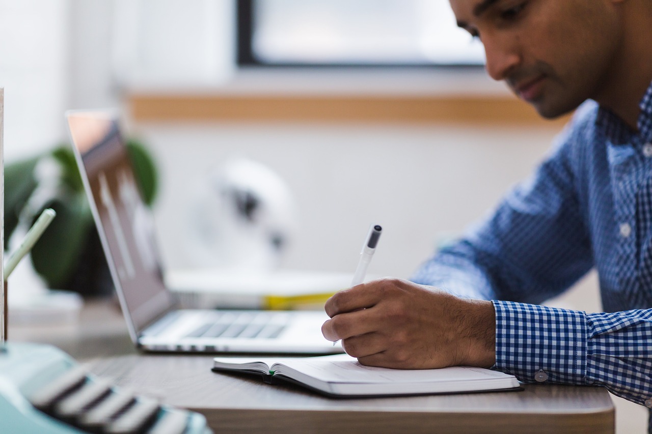 Photo of a man writing in a notepad next to a laptop computer.