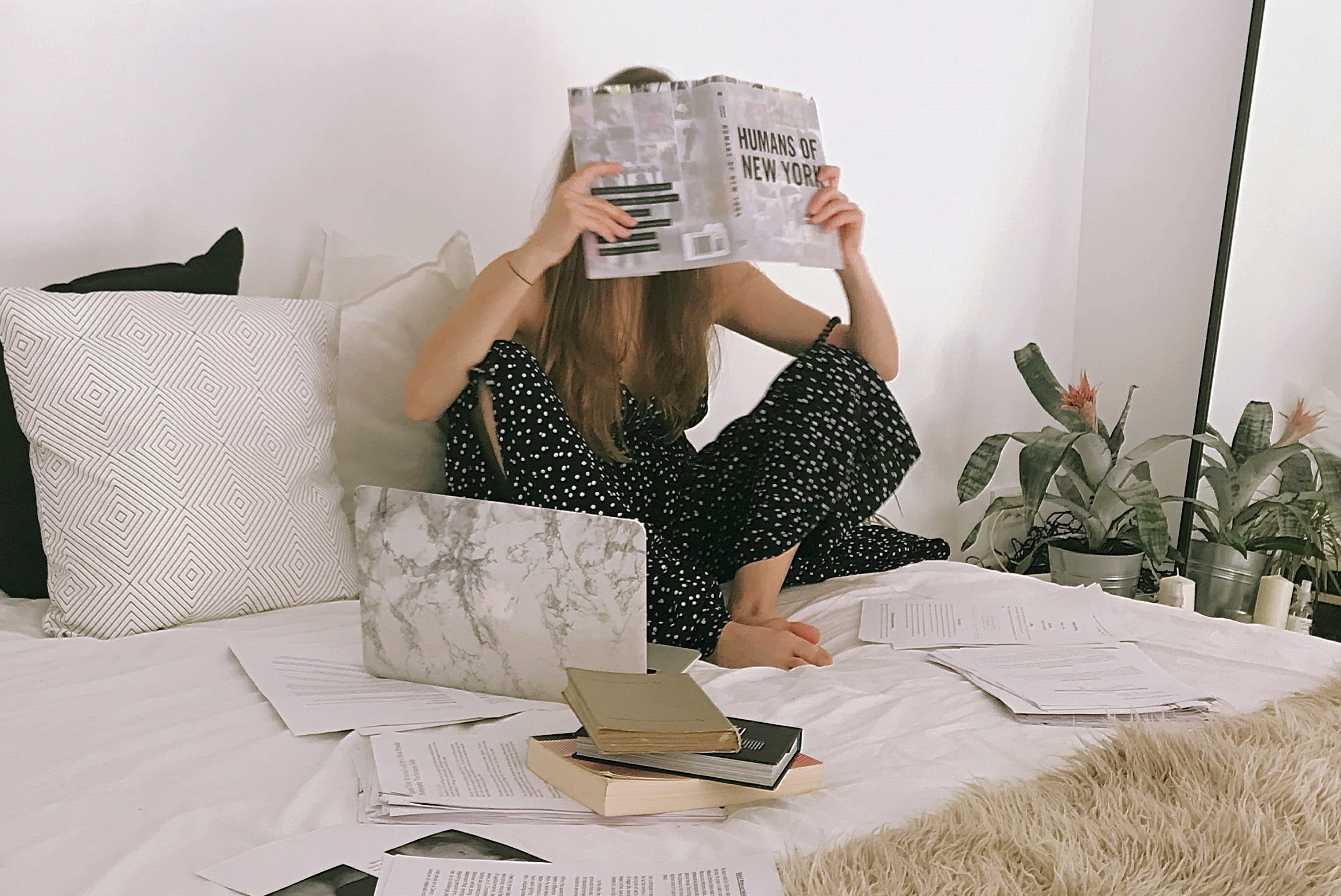 Photo of a woman sitting on a bed with a laptop, surrounded by books and papers.