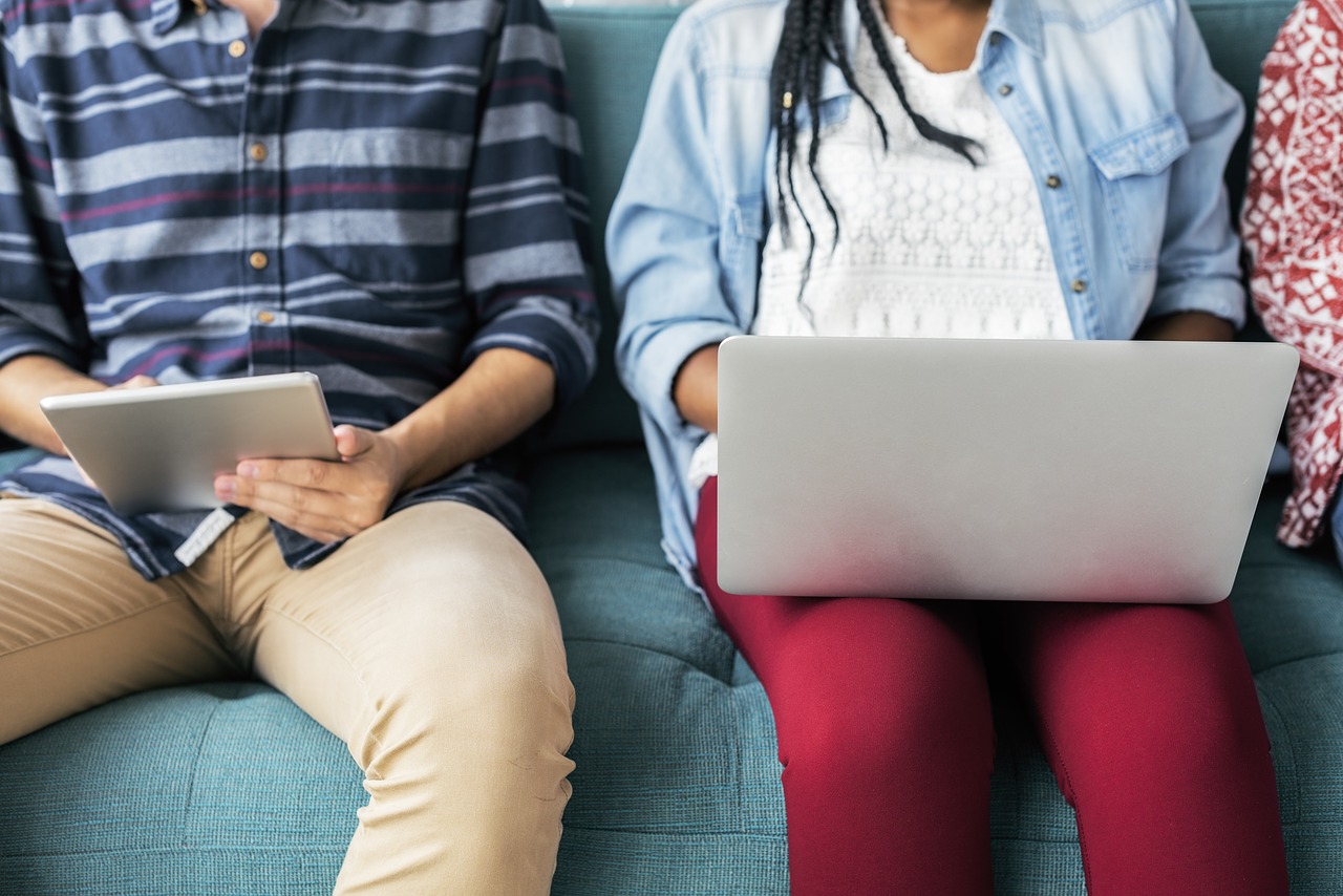 Photo of two people sitting on a couch, one with a tablet and the other with a laptop.