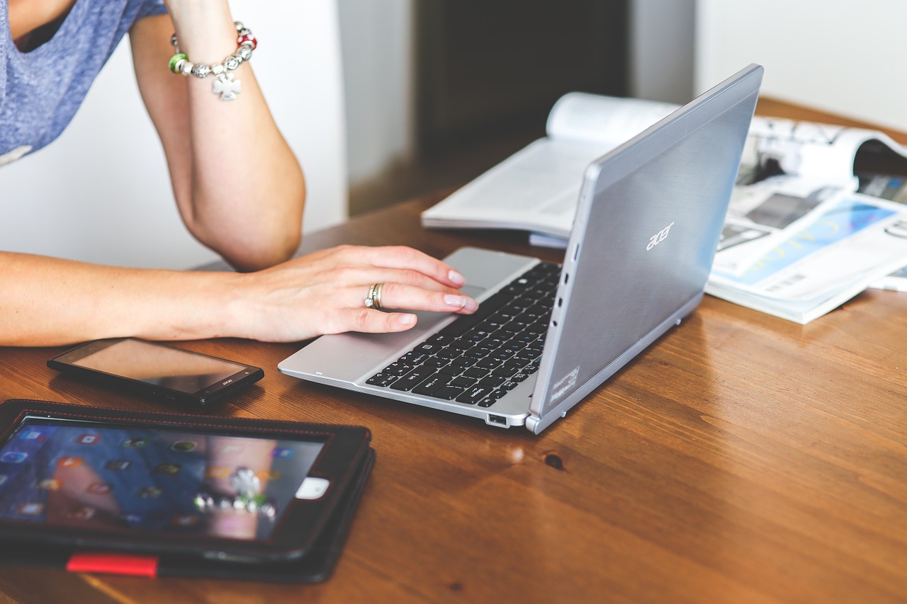 Photo of a woman working at a laptop with magazines and a tablet nearby.