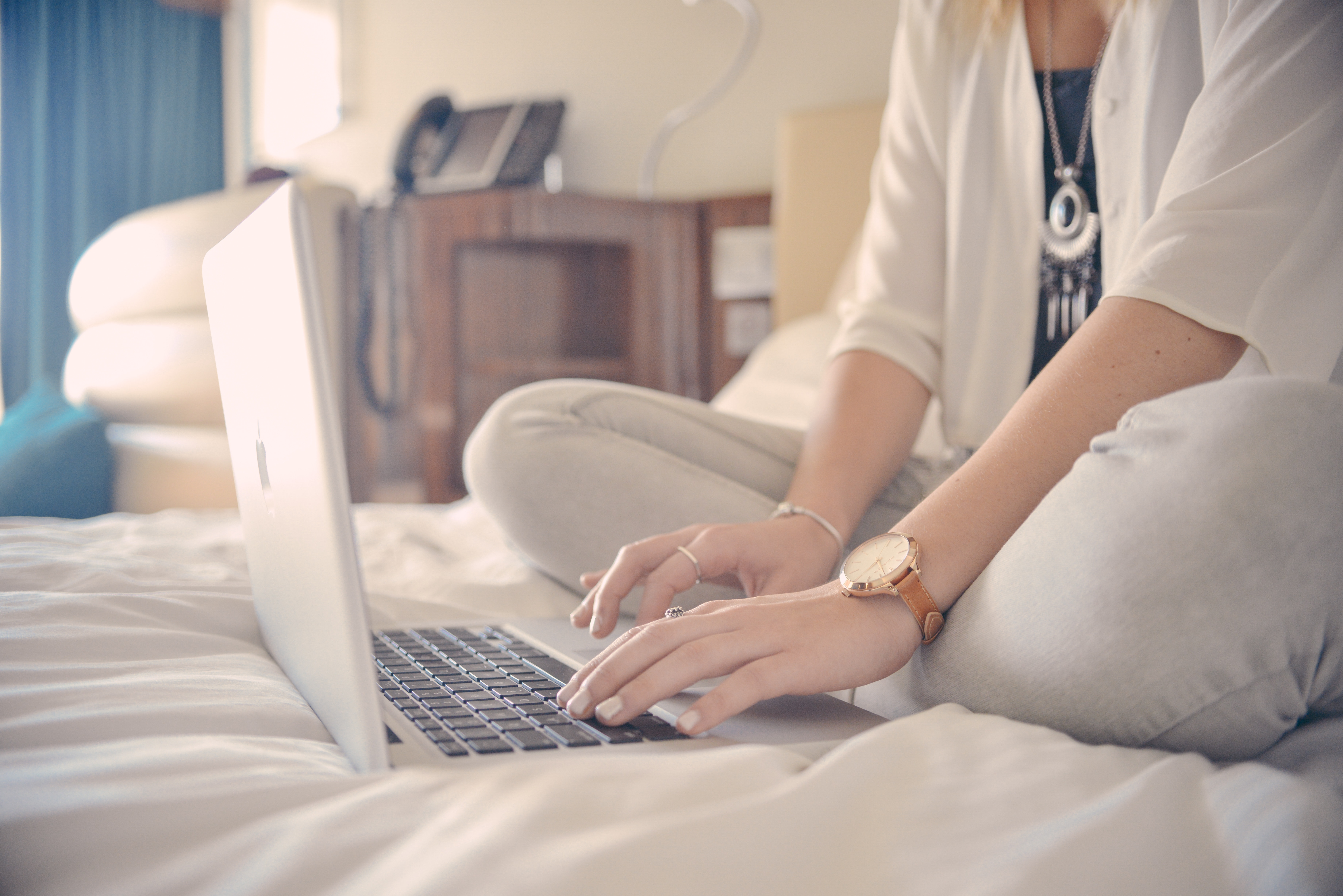 Photo of a woman sitting on a bed and typing on a laptop computer.